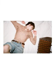 A young man wearing a net top and denim shorts lying on a white bed next to a patterned handbag