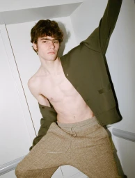 Shirtless male model wearing khaki trousers and an olive cardigan lifted over one arm against a white wall.