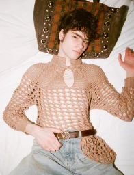 Young male model lying on a white surface wearing an open-weave beige knit top and denim jeans with a brown leather belt.