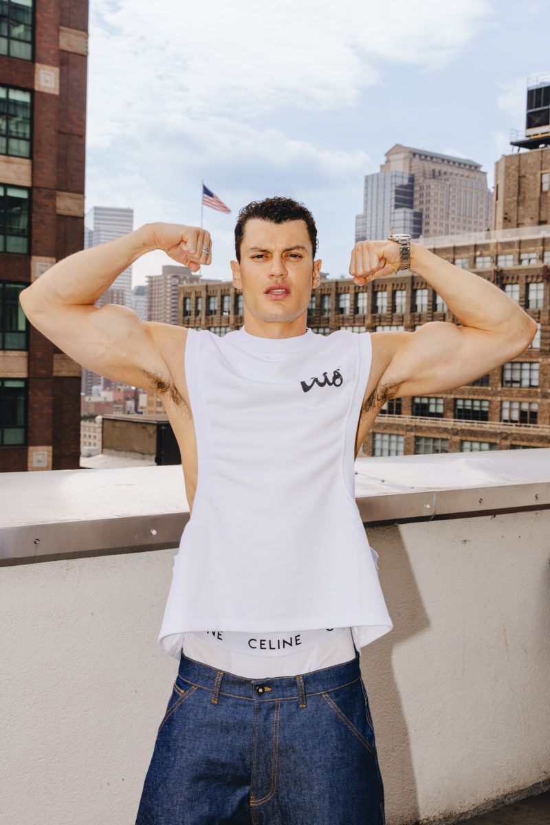 Fit male model flexing biceps on a city rooftop wearing a white tank top and blue jeans, with skyscrapers and the US flag in the background.