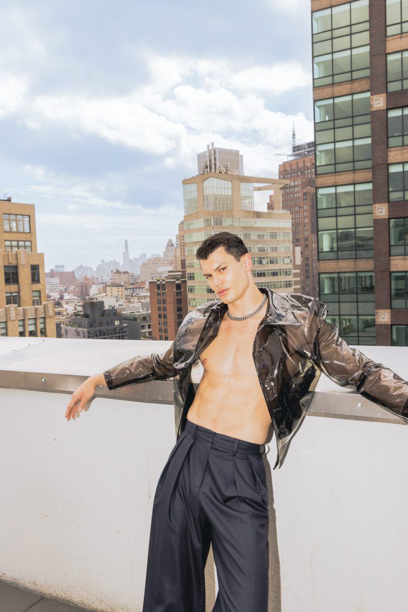 Young man poses on a rooftop with buildings in the background. He is wearing a see-through jacket and wide-legged black pants.