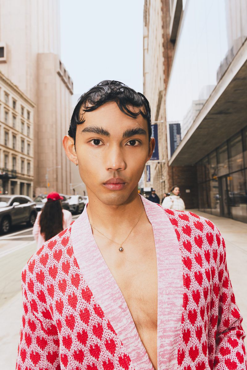 Close-up of a young adult man wearing a heart-patterned cardigan in an urban setting with buildings in the background on a city street.