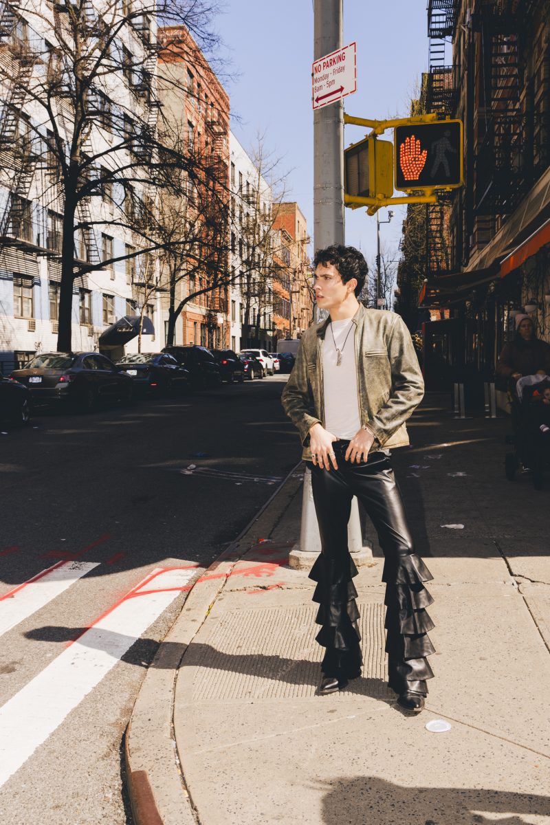 Adult male in a jacket and black ruffle pants standing on a sidewalk in a city with buildings and a traffic light in the background.
