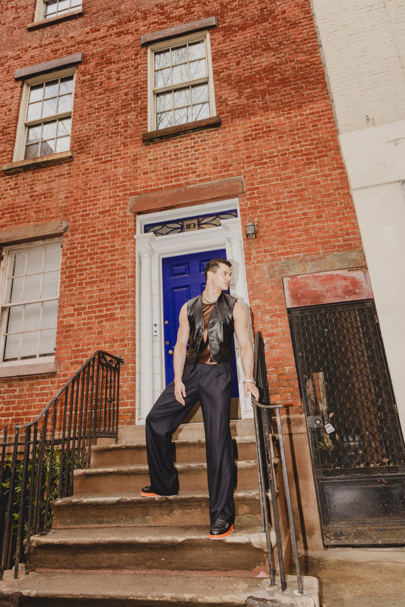 A well-built man in a black leather vest poses on brownstone steps with a blue door, brick facade, and black iron railings.