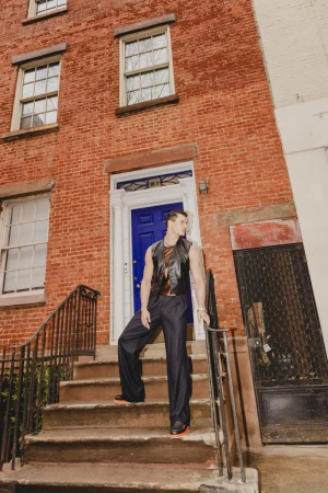 A well-built man in a black leather vest poses on brownstone steps with a blue door, brick facade, and black iron railings.