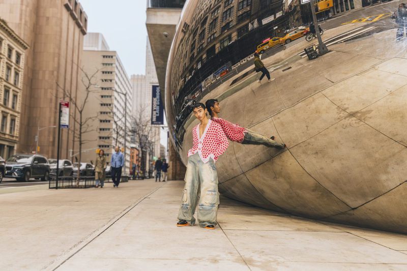 Young adult poses in front of shiny art installation in urban setting, wearing red patterned cardigan, distressed baggy denim and orange sneakers.