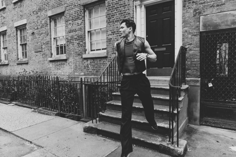 A man in a vest walks down stairs outside a brick building. Black and white photo features urban setting with steps, door, and railing.