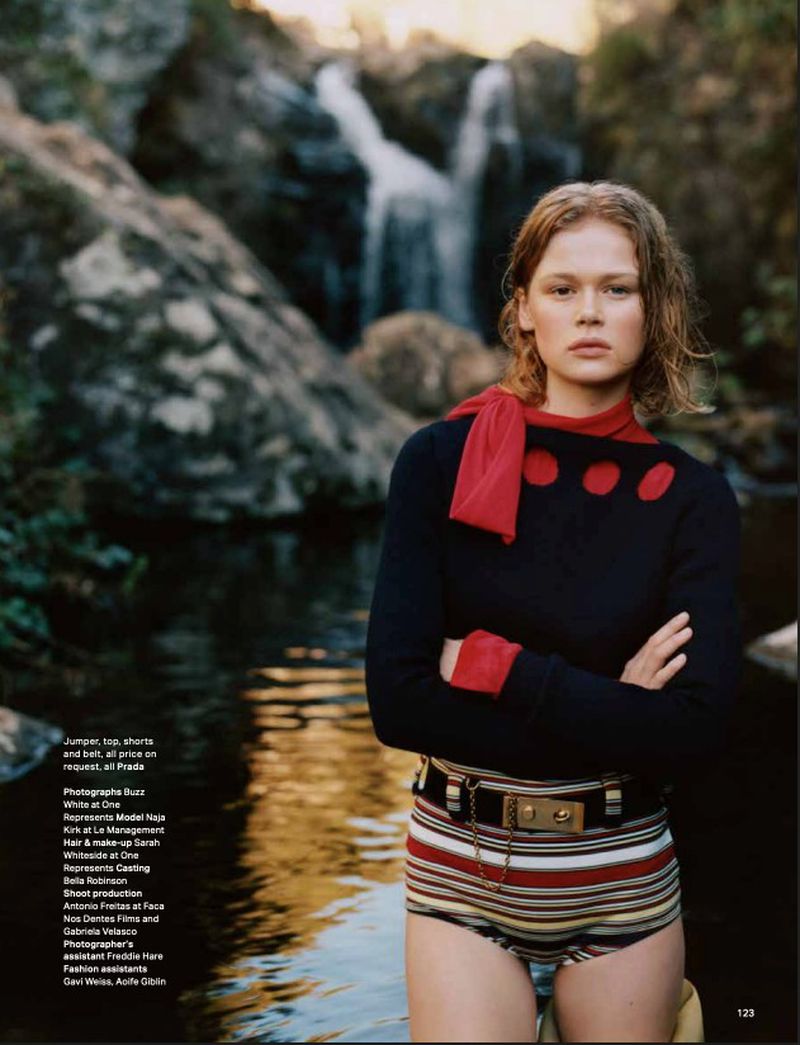 Young woman in a navy blue sweater and striped shorts stands in front of a waterfall in a lush outdoor setting. Fashion shot.