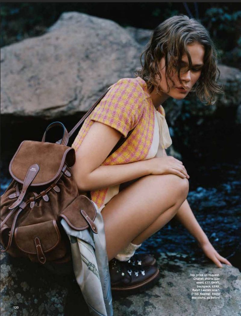 A woman in outdoor clothing is crouched by the water's edge with a backpack. She is surrounded by rocks in a natural setting.