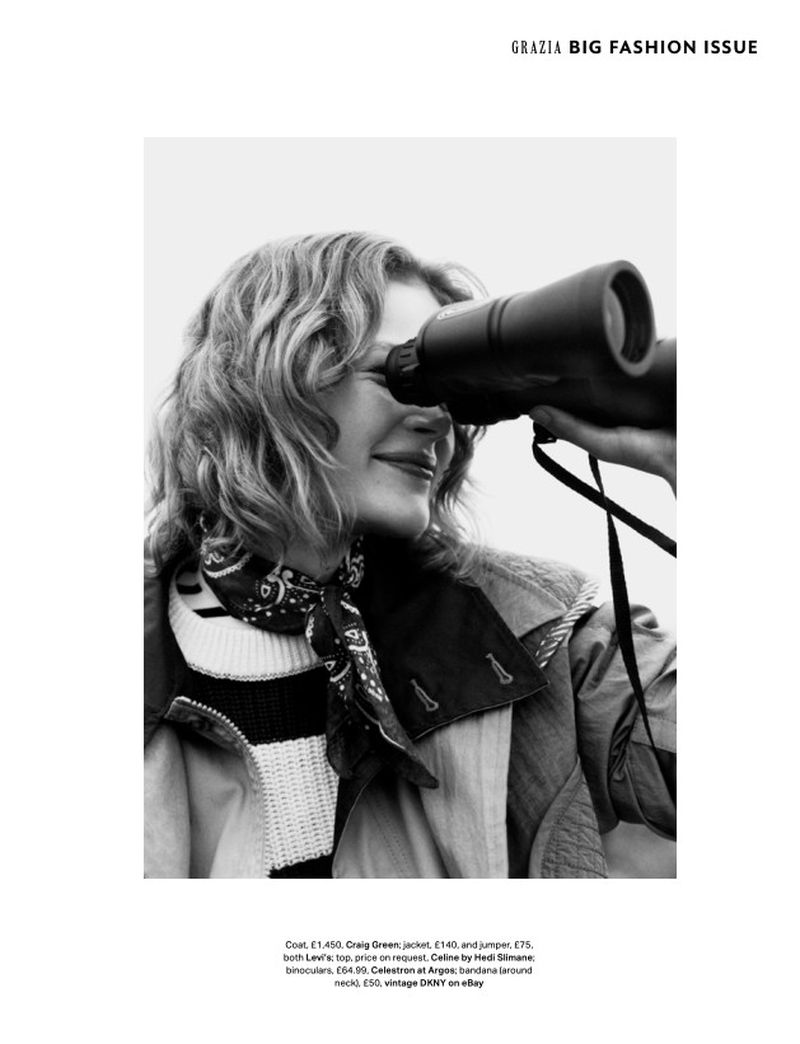 Black and white fashion shot of a curly-haired woman wearing a coat, sweater, and bandana, looking through binoculars, medium shot.