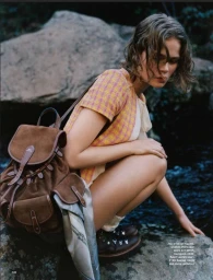 Model Naja Kirk crouching near water in a pink and yellow top with a brown suede backpack for Grazia UK