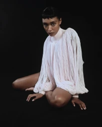 Model seated on a dark background wearing a textured white fringe garment with a high collar.