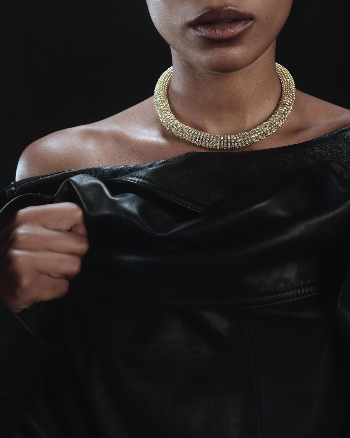 Close-up portrait of a model wearing a thick gold choker necklace and a dark leather top against a dark background.