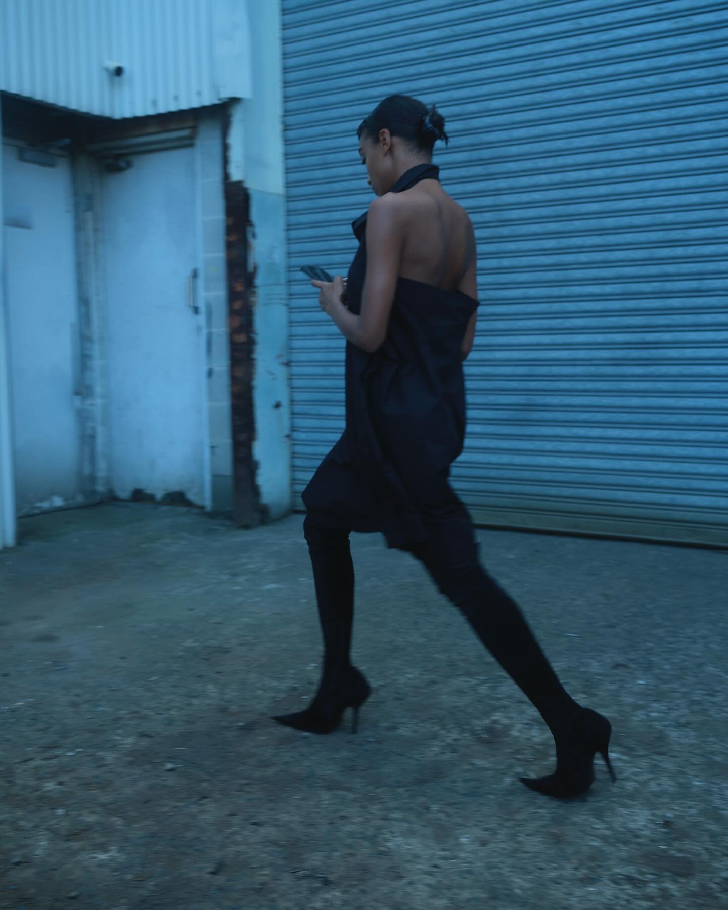 Woman in black dress and heels walks on street, checks smartphone, illuminated by cold blue light, shuttered door in background.