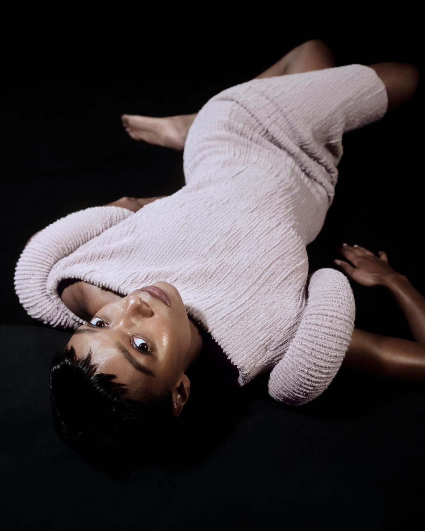 A woman with short black hair laying on a black surface wearing a textured knit dress, posing for a fashion or beauty editorial shoot.