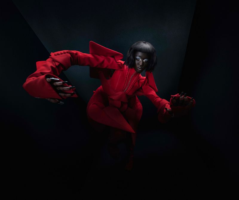 Woman with dark skin and a bob haircut poses in an avant-garde red outfit in a low-key fashion shot, emphasizing dramatic lighting and bold design.