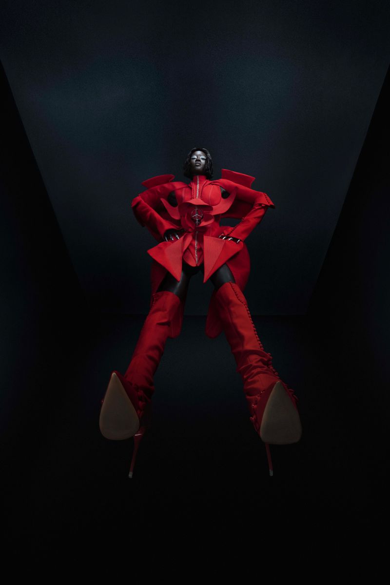 A person in a red avant-garde outfit poses dramatically, viewed from a low angle against a dark backdrop, highlighting high fashion.
