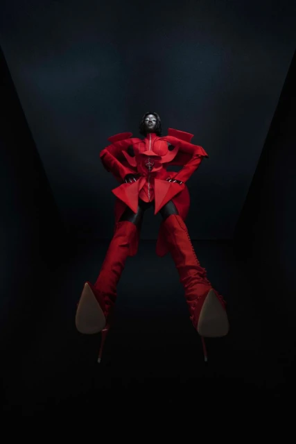 A person in a red avant-garde outfit poses dramatically, viewed from a low angle against a dark backdrop, highlighting high fashion.