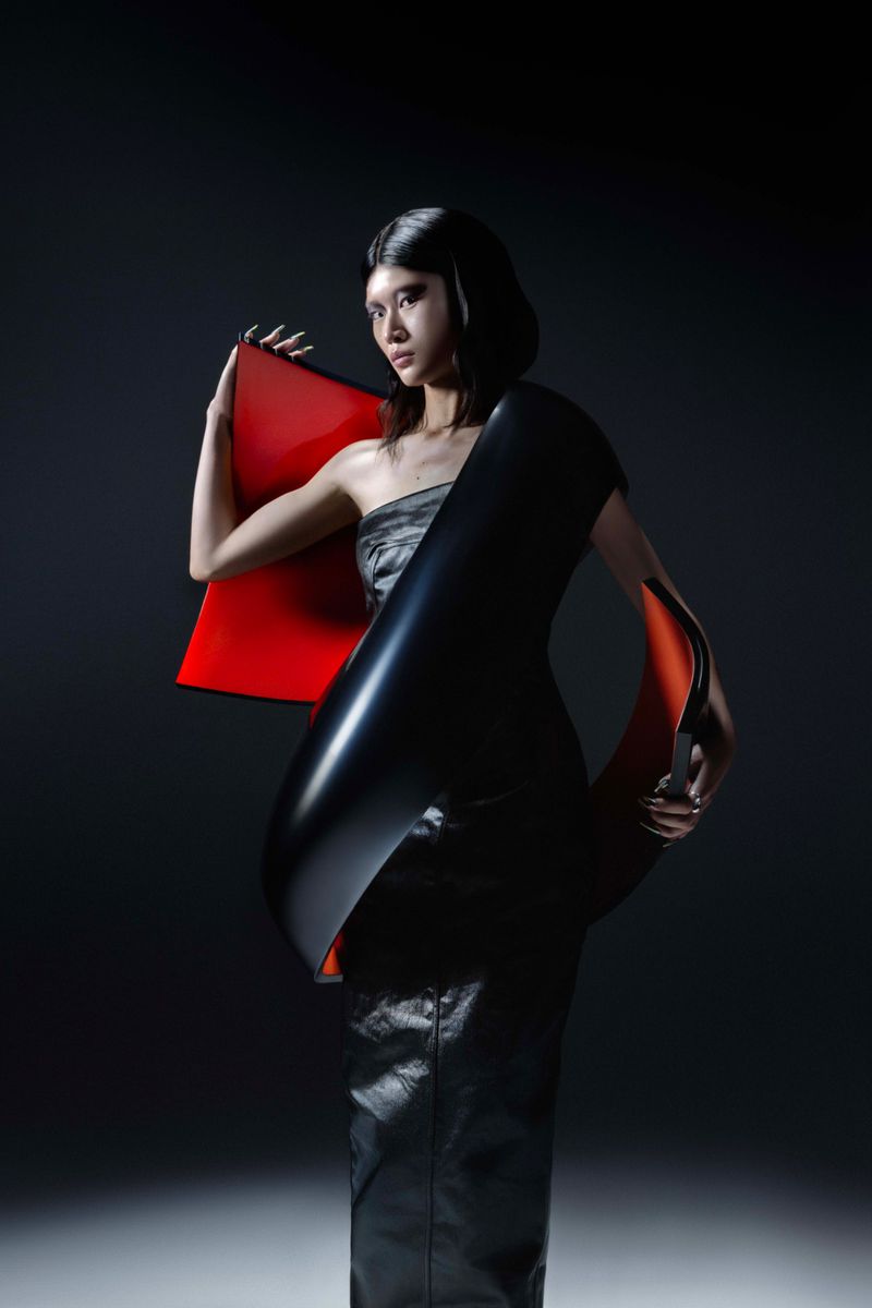 A female fashion model poses in a strapless grey top and black leather skirt, adorned with a black and red architectural accent piece.