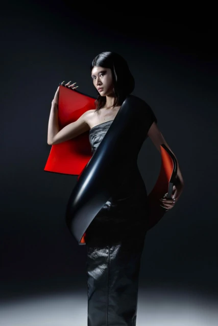 A female fashion model poses in a strapless grey top and black leather skirt, adorned with a black and red architectural accent piece.