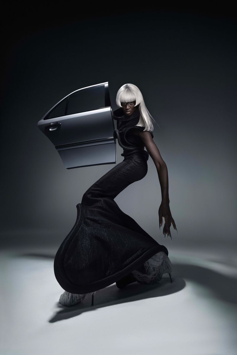 Artistic studio shot of a Black adult female in a black outfit and a white wig posing with a silver car door in a gray studio.