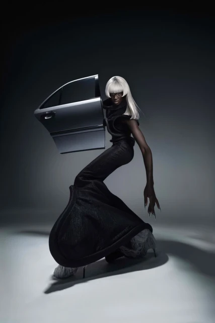 Artistic studio shot of a Black adult female in a black outfit and a white wig posing with a silver car door in a gray studio.