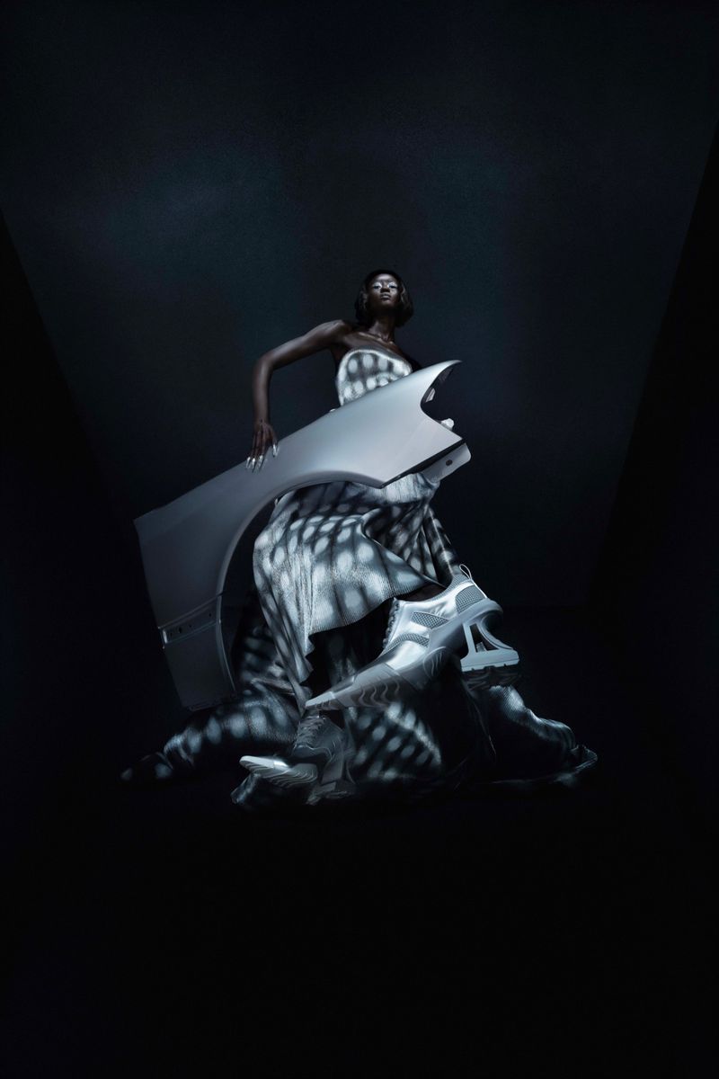 Fashion shot of an adult Black female posing with a gray auto body panel and shoes against a gradient dark-gray background.