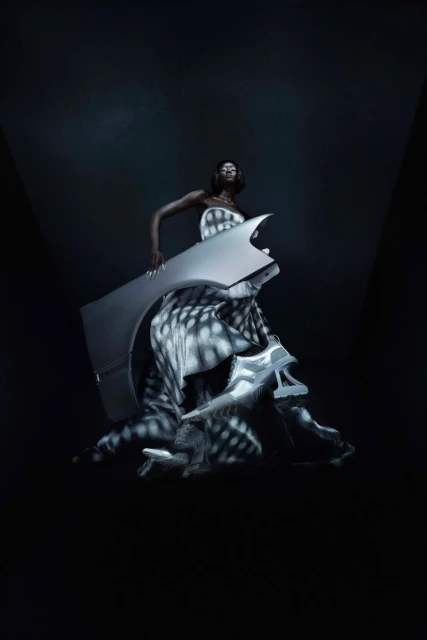 Fashion shot of an adult Black female posing with a gray auto body panel and shoes against a gradient dark-gray background.