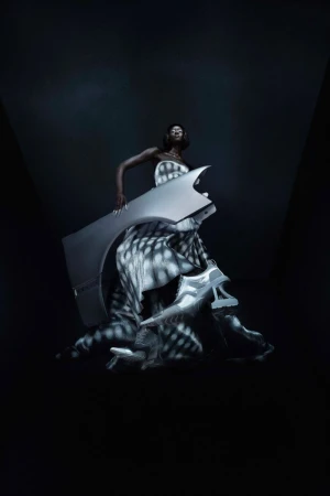Fashion shot of an adult Black female posing with a gray auto body panel and shoes against a gradient dark-gray background.