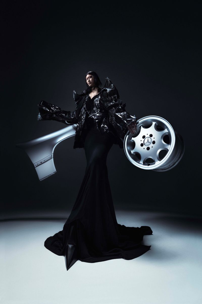Fashion photo of a woman in black sculptural dress posing with automotive parts and wheel against dark backdrop in studio setting.