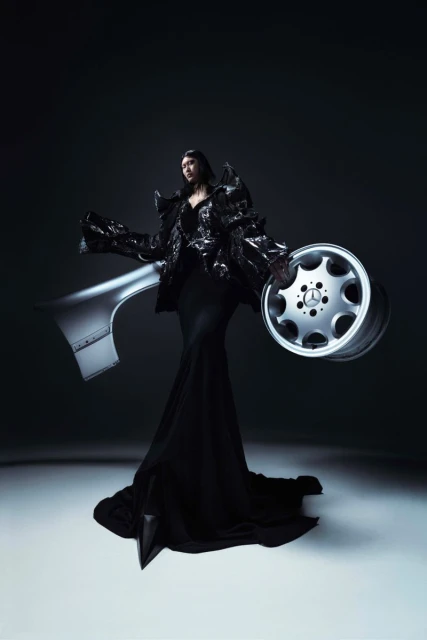 Fashion photo of a woman in black sculptural dress posing with automotive parts and wheel against dark backdrop in studio setting.