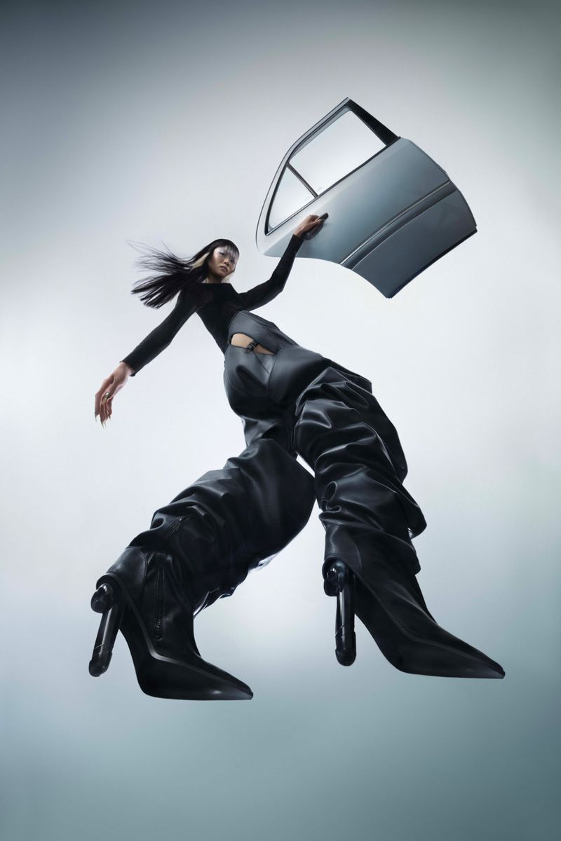 Fashion-forward image featuring a woman posing with a car door, wearing black clothing and oversized black heeled boots against a gradient grey background.