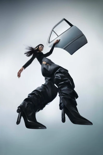 Fashion-forward image featuring a woman posing with a car door, wearing black clothing and oversized black heeled boots against a gradient grey background.