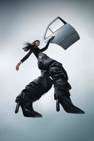 Fashion-forward image featuring a woman posing with a car door, wearing black clothing and oversized black heeled boots against a gradient grey background.