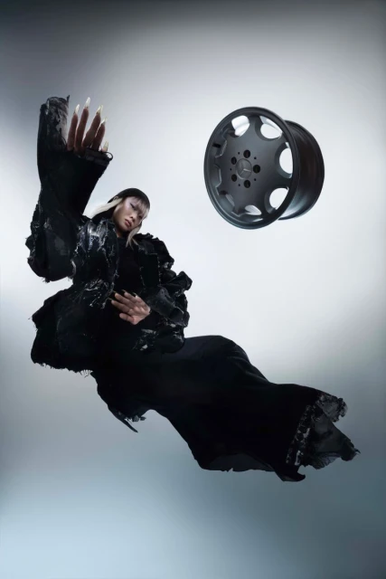 Woman in couture gown poses next to a car wheel against a gradient grey background in a fashion editorial with theatrical costume details.