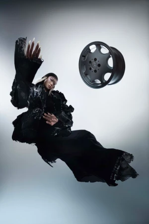 Woman in couture gown poses next to a car wheel against a gradient grey background in a fashion editorial with theatrical costume details.