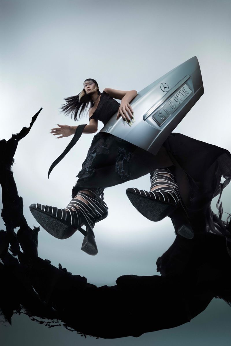 Fashionable Asian woman poses with a Mercedes car part, wearing a black dress and patterned shoes in a studio setting with soft lighting.