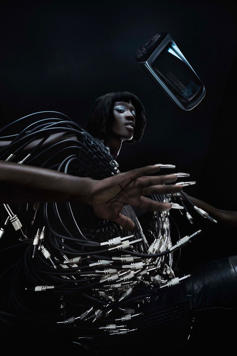 A woman with dark skin and short black hair poses with audio cables while a clear rectangular perfume bottle floats above her.