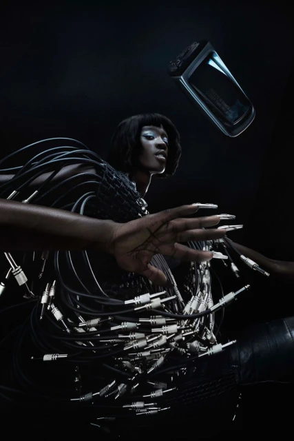 A woman with dark skin and short black hair poses with audio cables while a clear rectangular perfume bottle floats above her.