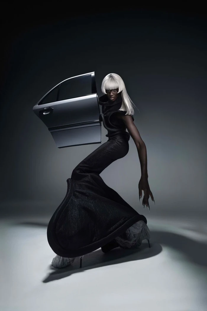 Model with platinum blonde wig wearing black gown beside car door in studio