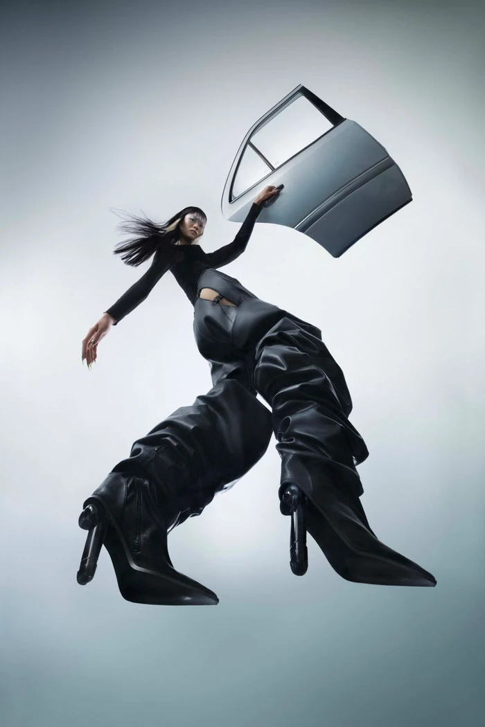 Model with dark hair wearing black garments and high boots holding a car door in a low-angle fashion editorial shot