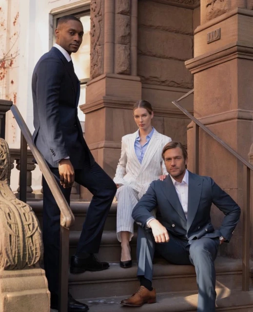 Three people in suits pose on the steps of a brownstone building. An adult male stands while an adult female and adult male sit.