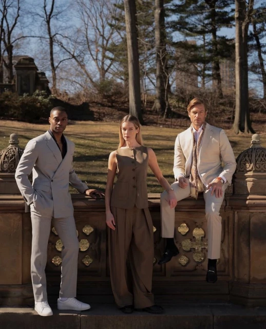 Three fashion models in neutral-tone suits pose on a stone ledge in a park setting with bare trees and a building visible in the background.