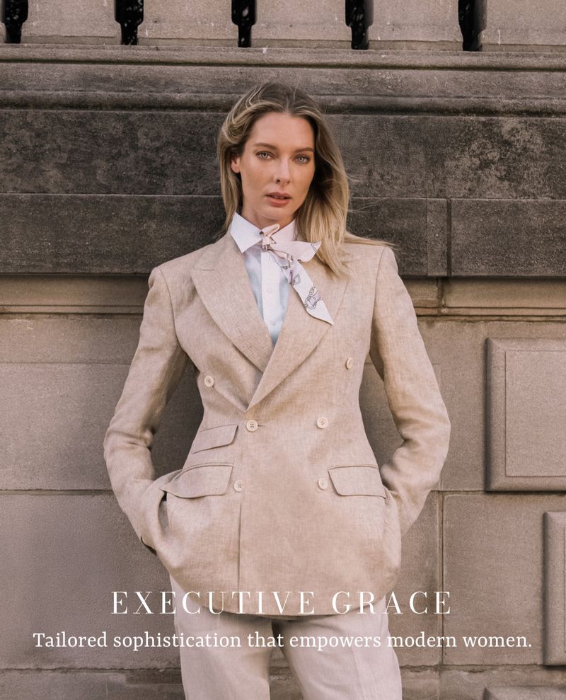 Blonde woman in light tan blazer and pants poses against a stone wall with black metal accents, looking confidently at the camera.