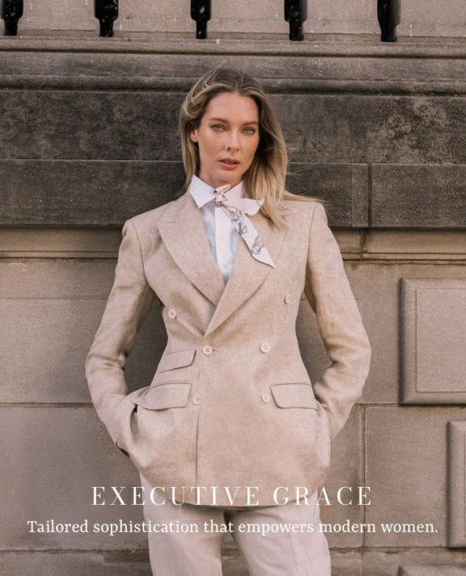 Blonde woman in light tan blazer and pants poses against a stone wall with black metal accents, looking confidently at the camera.