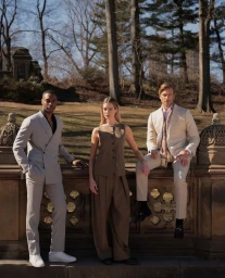 Three fashion models posing outdoors leaning against an ornate stone balustrade in a park setting.