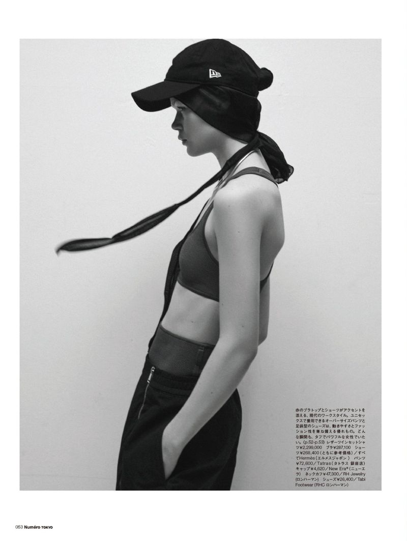 Black and white fashion shot of a young adult woman in a cap, scarf, bra top, and overalls against a seamless white background.