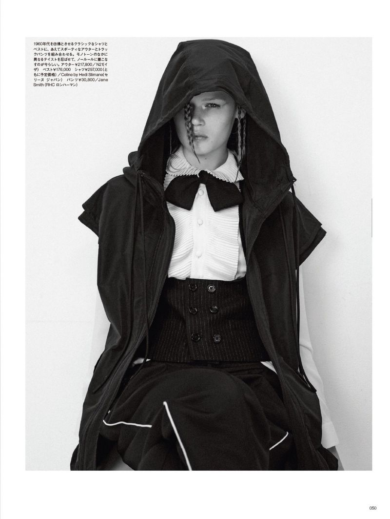 High-fashion black-and-white portrait of a model in a hooded jacket, vest, pleated shirt, and skirt, posing indoors in front of a stark white wall.