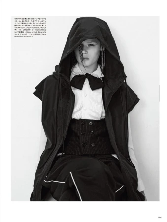 High-fashion black-and-white portrait of a model in a hooded jacket, vest, pleated shirt, and skirt, posing indoors in front of a stark white wall.