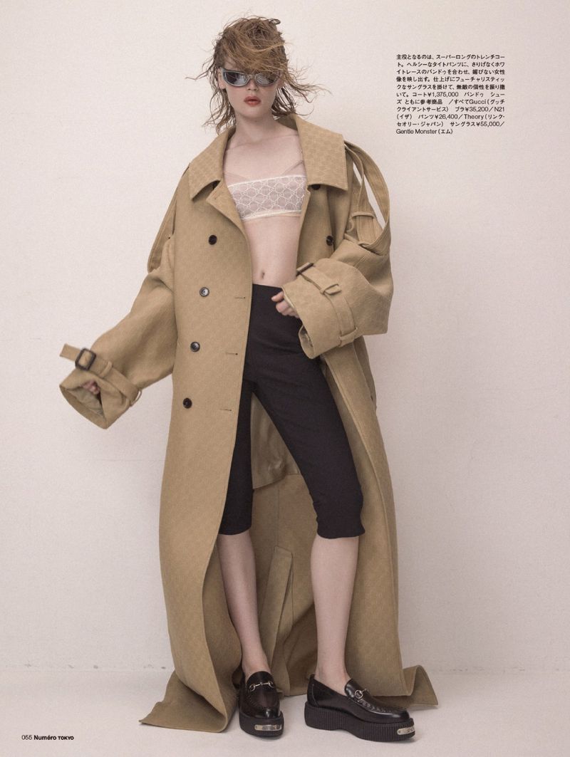 Fashion editorial shot of a female model in a beige oversized trench coat, bra top, black shorts, and chunky platform loafers in a photo studio.